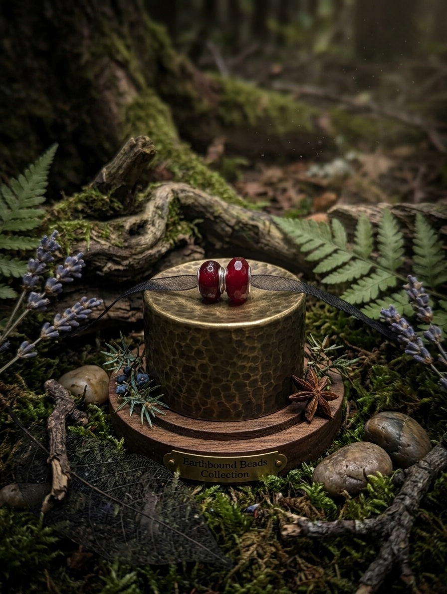 Two deep red resin European-style charm beads with silver cores displayed on a gold hammered pedestal stand labelled 'Earthbound Beads Collection', surrounded by moss, ferns, lavender, and star anise in a forest setting