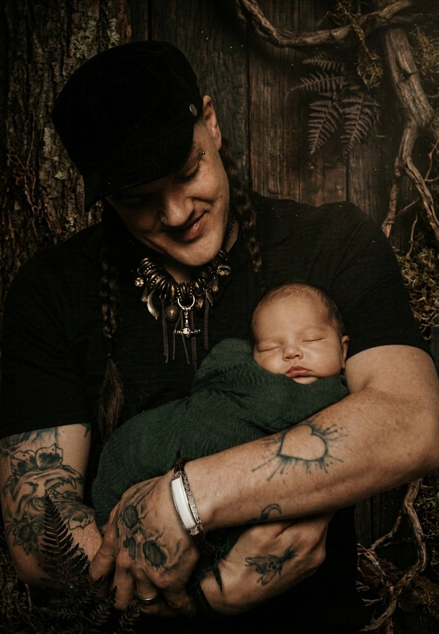 Tattooed man wearing the Nordic Spirit bracelet cradling a sleeping newborn in a dark, moody forest-inspired setting