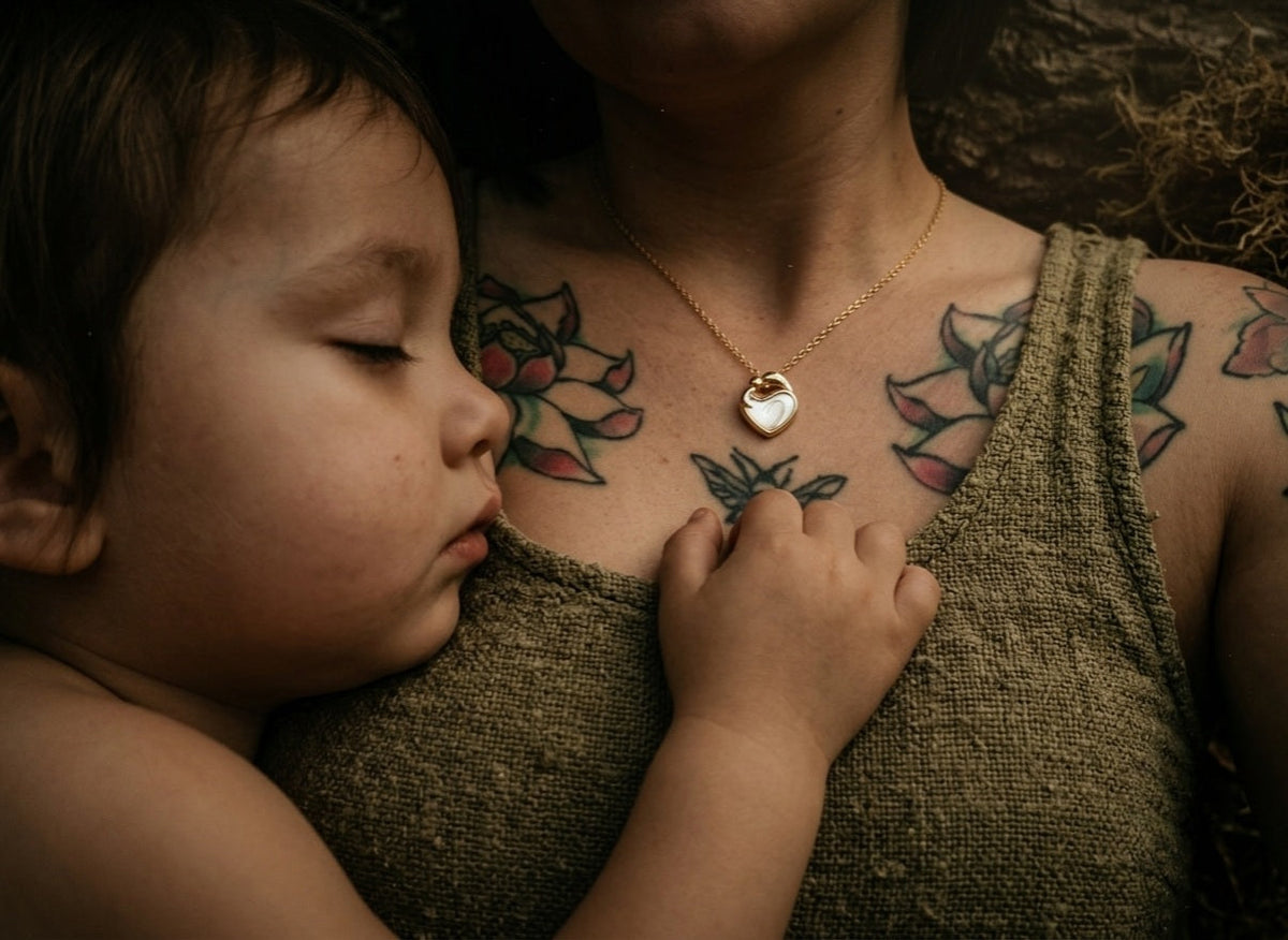 Tattooed mother wearing a gold breast milk pendant necklace cradling her sleeping toddler against her chest