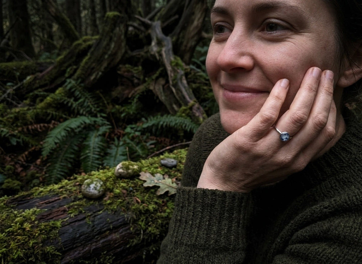 Smiling woman wearing the willow ring with a blue foils, resting her hand against her cheek in a lush forest setting