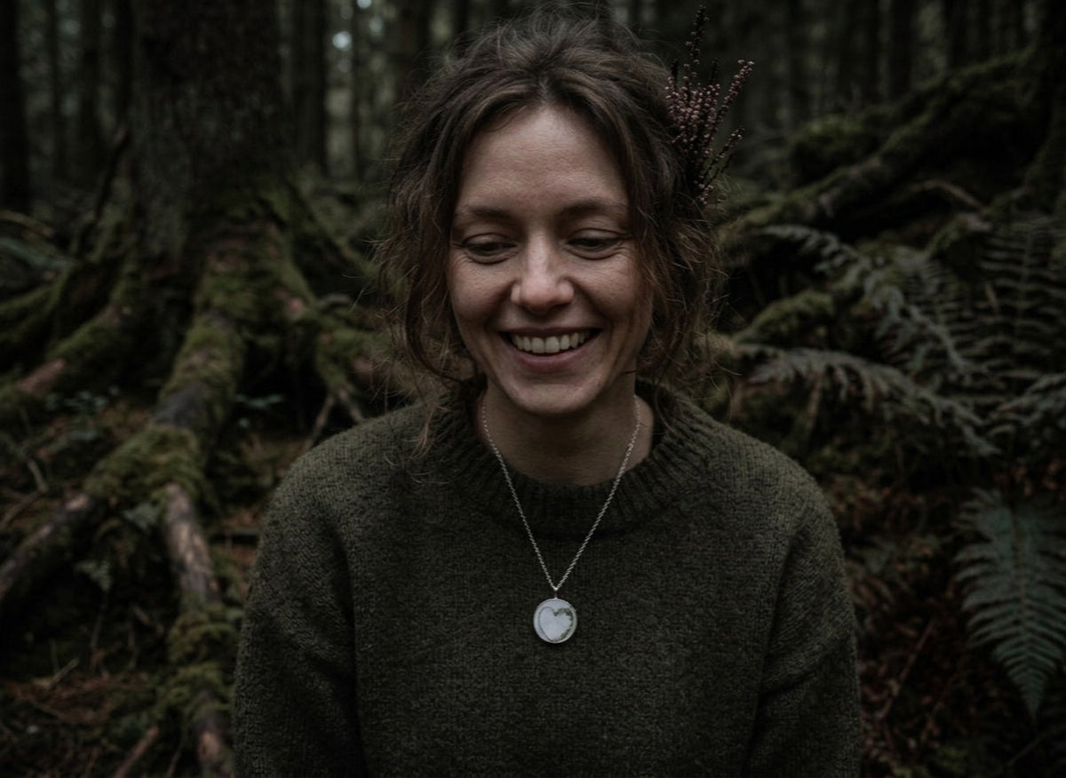A circular pendant hung over a wooden log with blurred foliage in the background. The pendant has a breastmilk stone with a baby hair heart in it with green flakes.