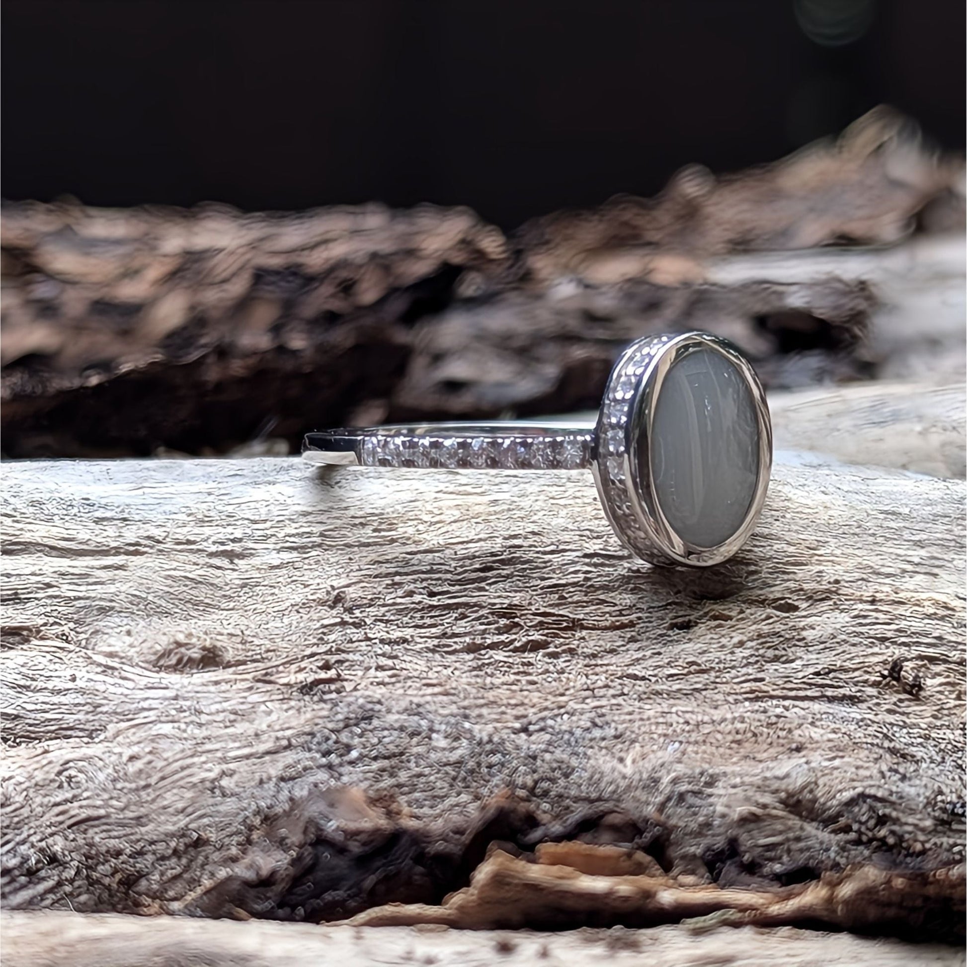 This image features a sterling silver ring with an oval-shaped milky white cabochon, breastmilk and resin, set on a band adorned with small clear stones. The ring is displayed on a piece of weathered wood. A beautiful breastmilk ring with sterling silver and clear stones. Made by Milky Way Spirit & Soul Keepsakes.