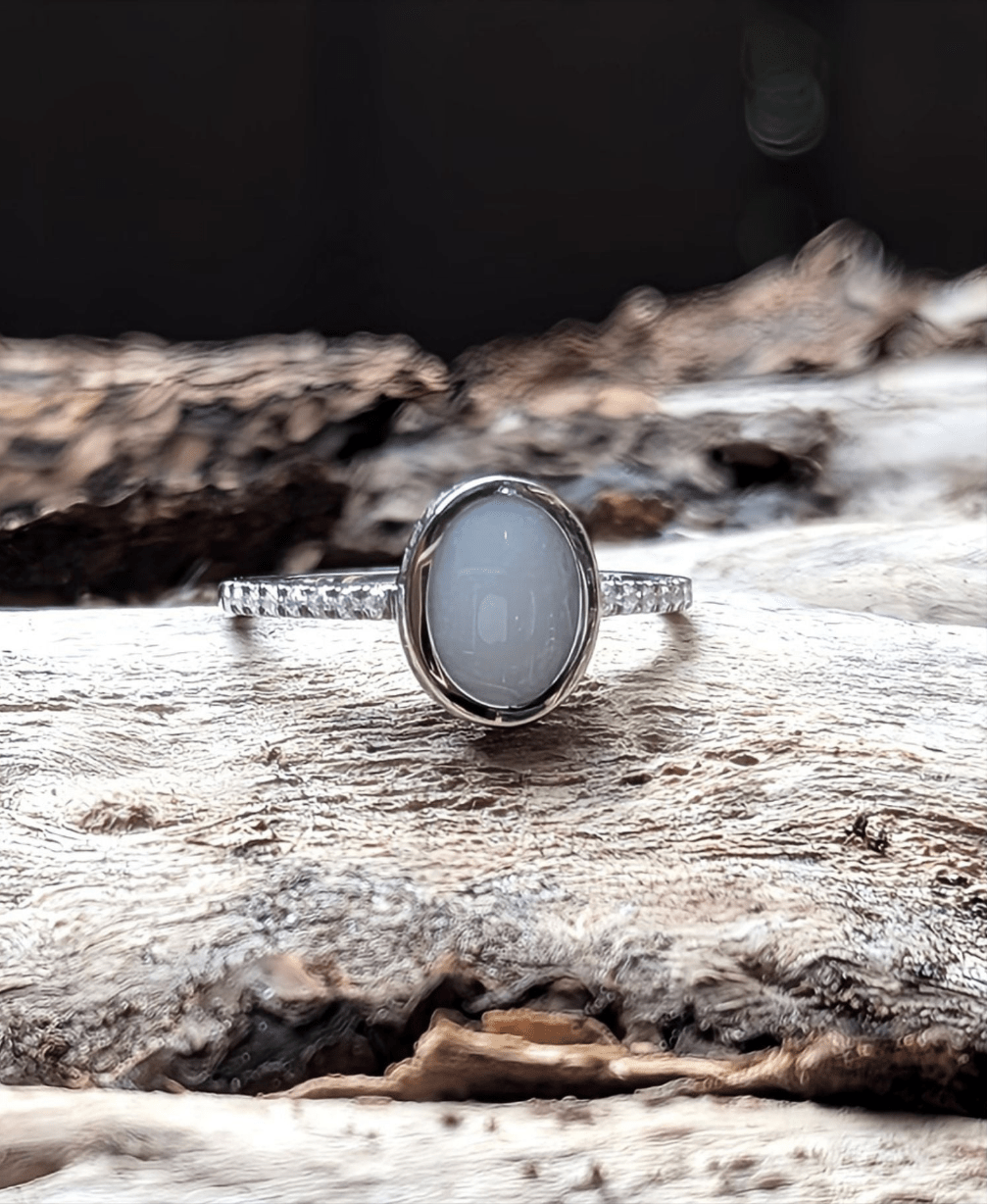 This image features a sterling silver ring with an oval-shaped milky white cabochon, breastmilk and resin, set on a band adorned with small clear stones. The ring is displayed on a piece of weathered wood. A beautiful breastmilk ring with sterling silver and clear stones. Made by Milky Way Spirit & Soul Keepsakes.