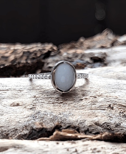This image features a sterling silver ring with an oval-shaped milky white cabochon, breastmilk and resin, set on a band adorned with small clear stones. The ring is displayed on a piece of weathered wood. A beautiful breastmilk ring with sterling silver and clear stones. Made by Milky Way Spirit & Soul Keepsakes.