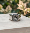 Silver ring with a square stone holding ash on a wooden surface with a blurred natural background.