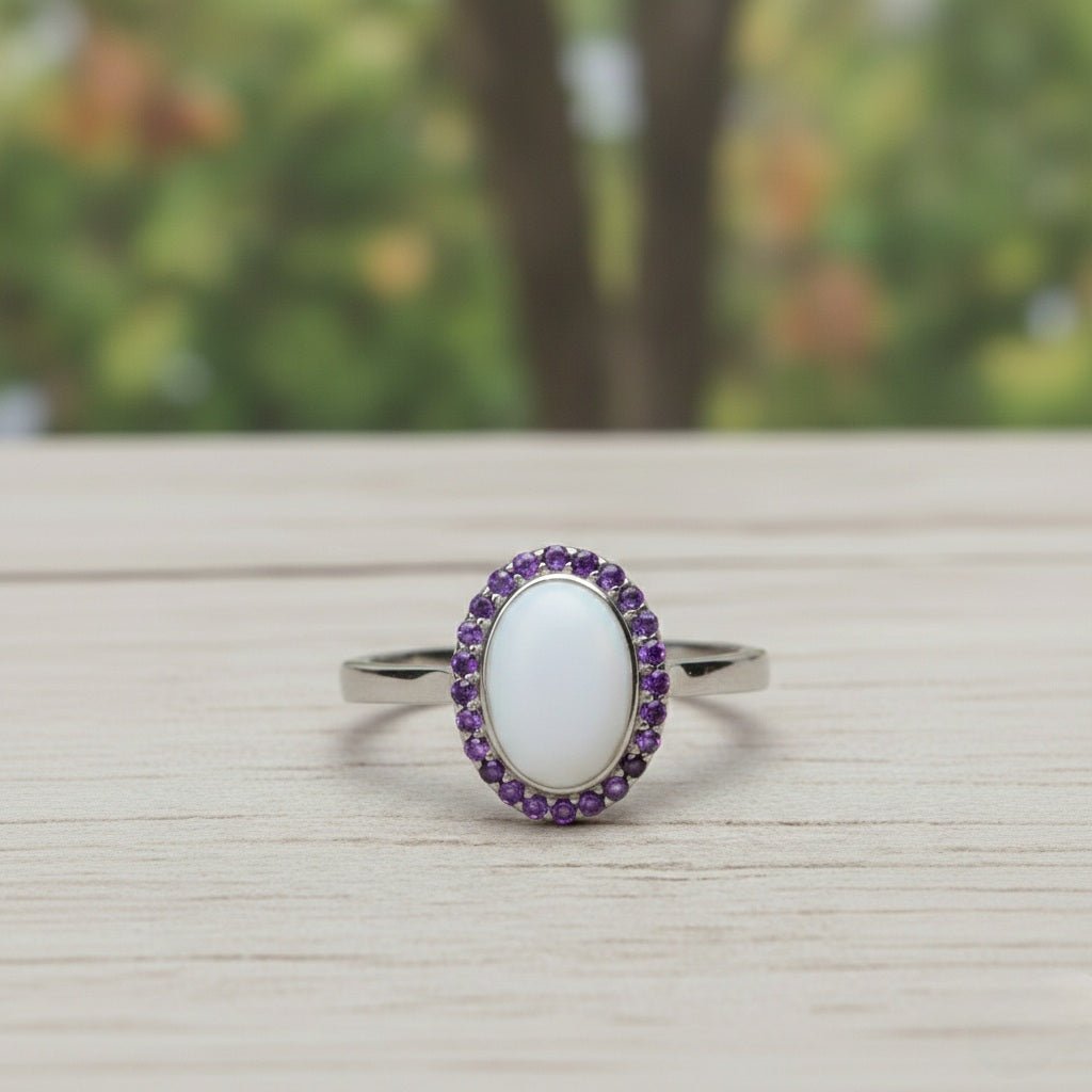 An elegant silver ring featuring a large, oval, smooth white stone centerpiece, encircled by a halo of small, vibrant purple amethyst crystals. The ring is displayed at a slight angle on a light-grained wooden surface with a soft-focus forest background.