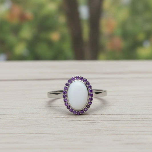 An elegant silver ring featuring a large, oval, smooth white stone centerpiece, encircled by a halo of small, vibrant purple amethyst crystals. The ring is displayed at a slight angle on a light-grained wooden surface with a soft-focus forest background.