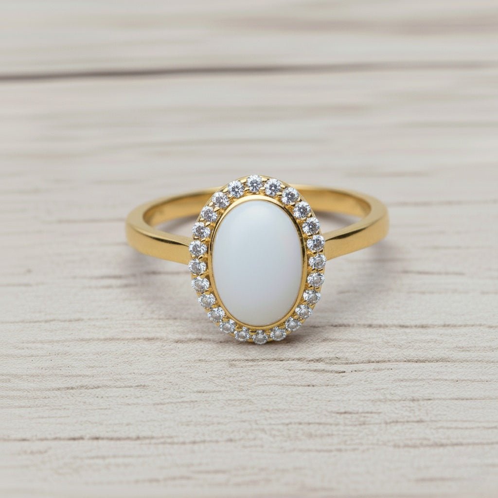 Milky Moments - Milky Way Spirit & Soul Keepsakes. An elegant gold ring featuring a large, oval, smooth white stone centerpiece, encircled by a halo of small, clear crystals. The ring is displayed at a slight angle on a light-grained wooden surface.