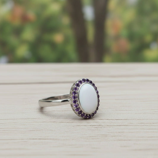 An elegant silver ring featuring a large, oval, smooth white stone centerpiece, encircled by a halo of small, vibrant purple amethyst crystals. The ring is displayed at a slight angle on a light-grained wooden surface with a soft-focus forest background.